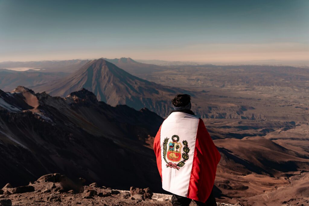 Man stands on Andean peak wrapped in Peru flag, overlooking stunning landscape.